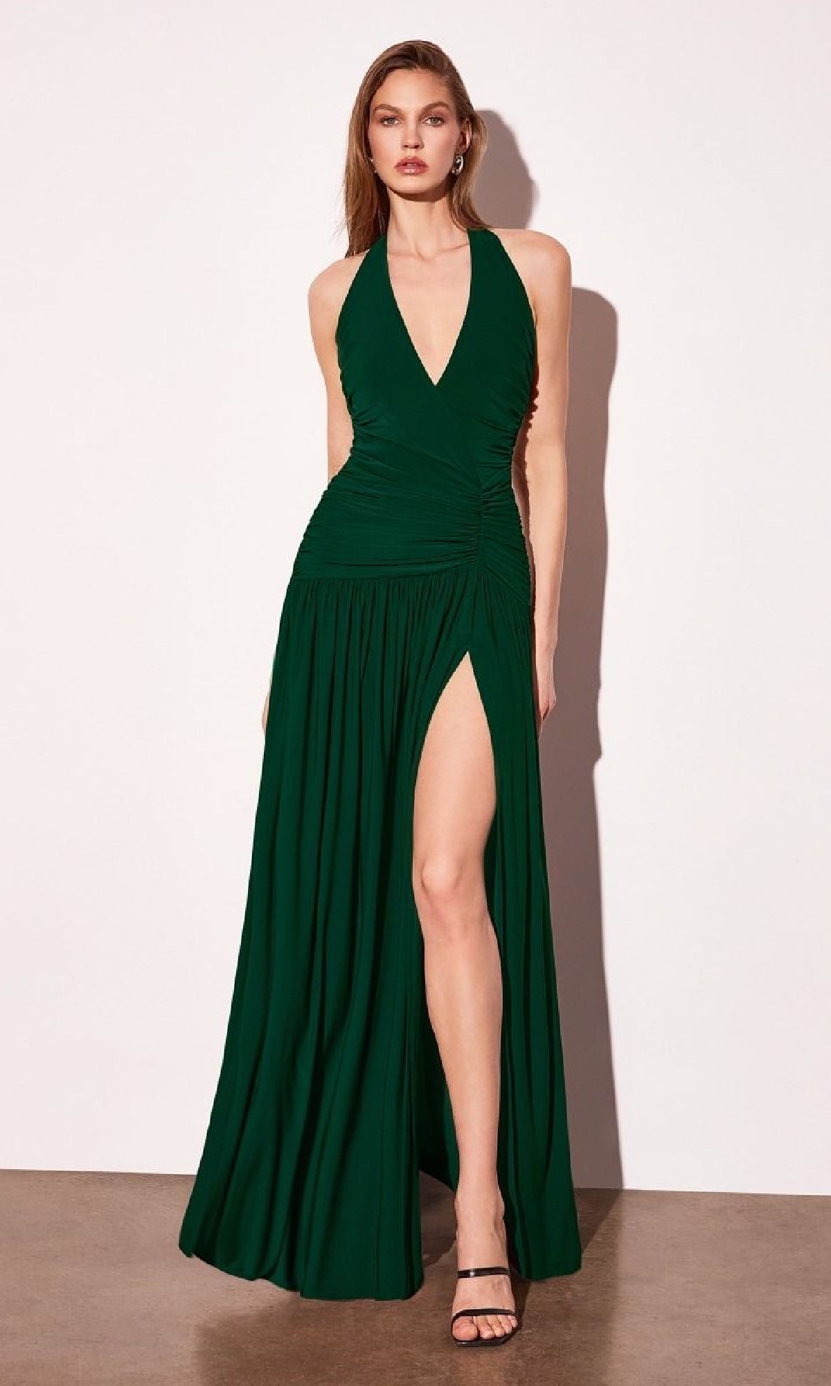 A woman poses against a white wall and concrete floor, wearing the Amoris T502 Backless Halter Prom Dress with Slit—a deep green, backless gown featuring a high thigh slit for a stunning, standout look.